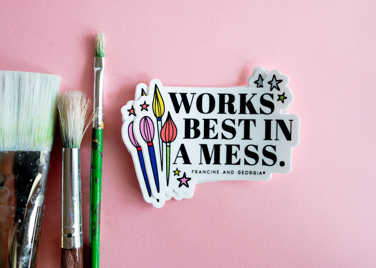 The works best in a mess sticker on a pink background next to paint brushes.
