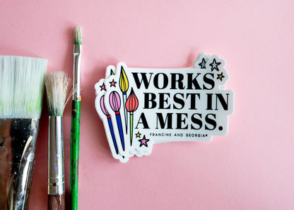 The works best in a mess sticker on a pink background next to paint brushes.